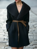 Black Lumina Wear Tabitha shawl-collar wrap coat with faux leather belt, shown on a model in a city setting.