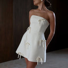 White satin strapless mini dress with bow accents by Lumina Vancouver, featuring a fitted bodice and elegant, sleek design.